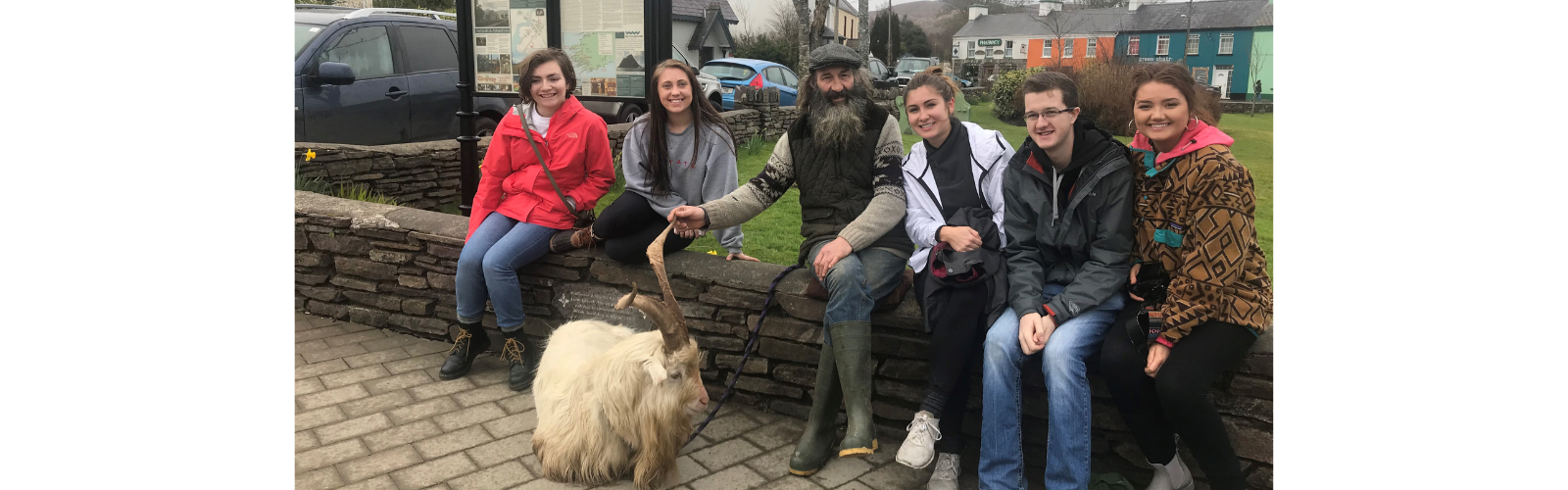 Five students sit next to a man with a beard holding onto a fluffy white goat with tall horns. 